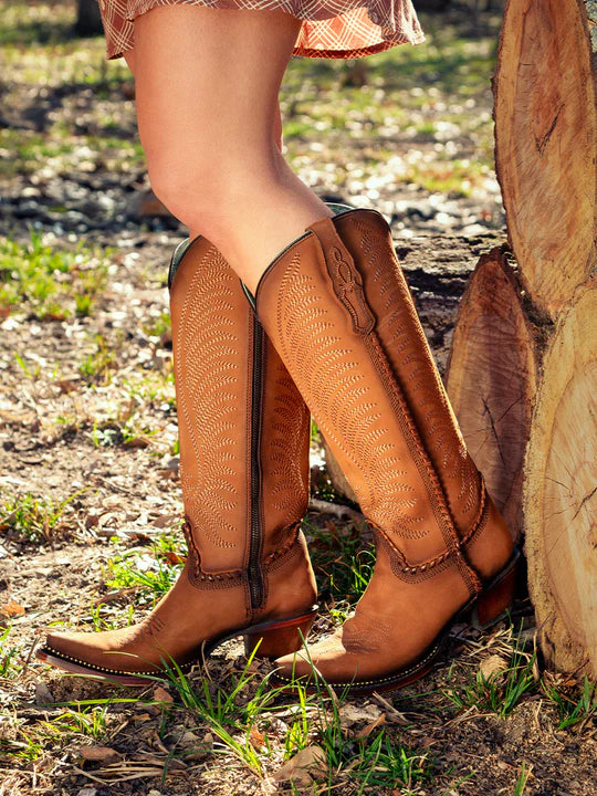 Brown cowboy boots worn by a person standing near wooden logs outdoors.