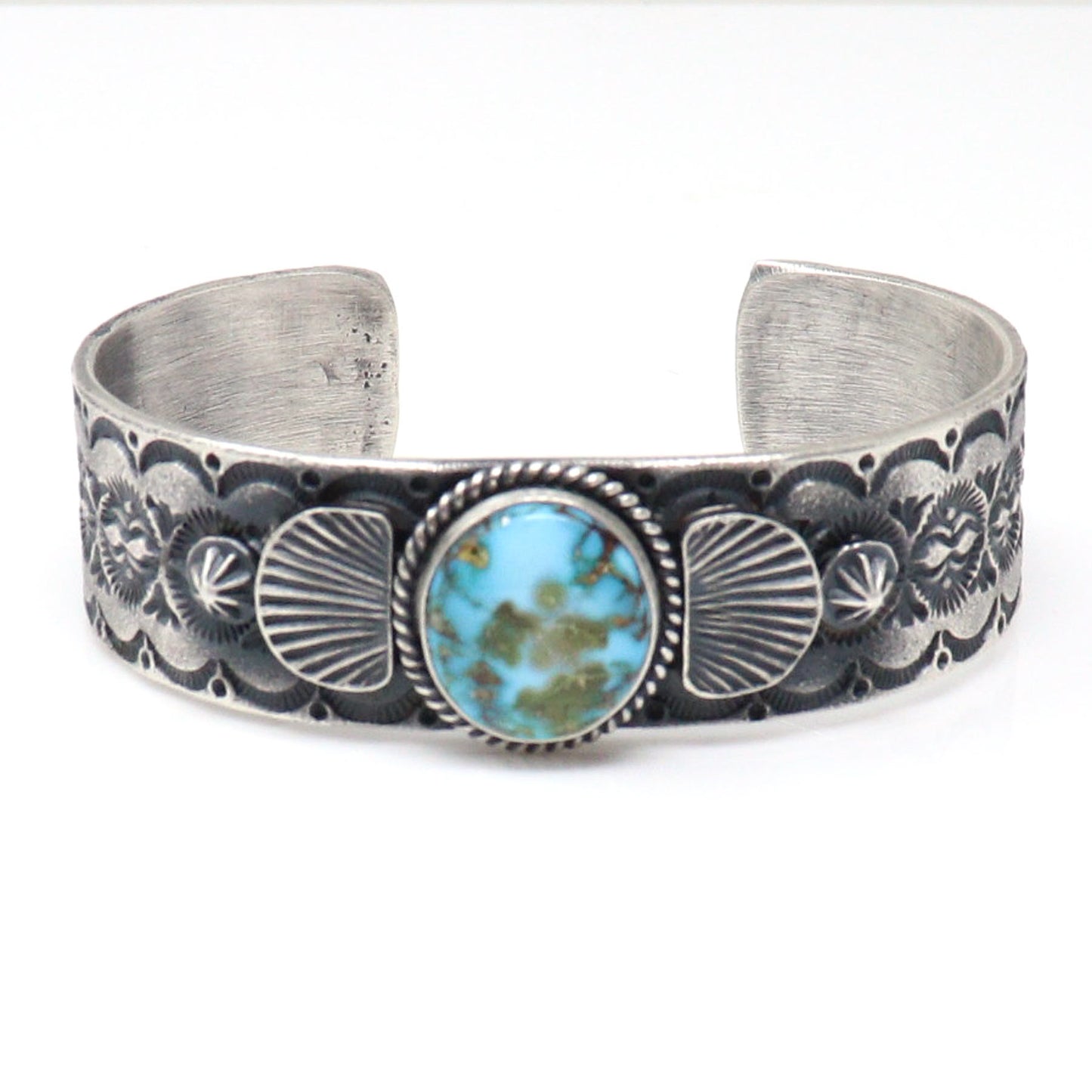 A sterling silver cuff bracelet featuring a central oval turquoise stone with natural matrix, surrounded by detailed floral and fan-shaped engravings on a white background.