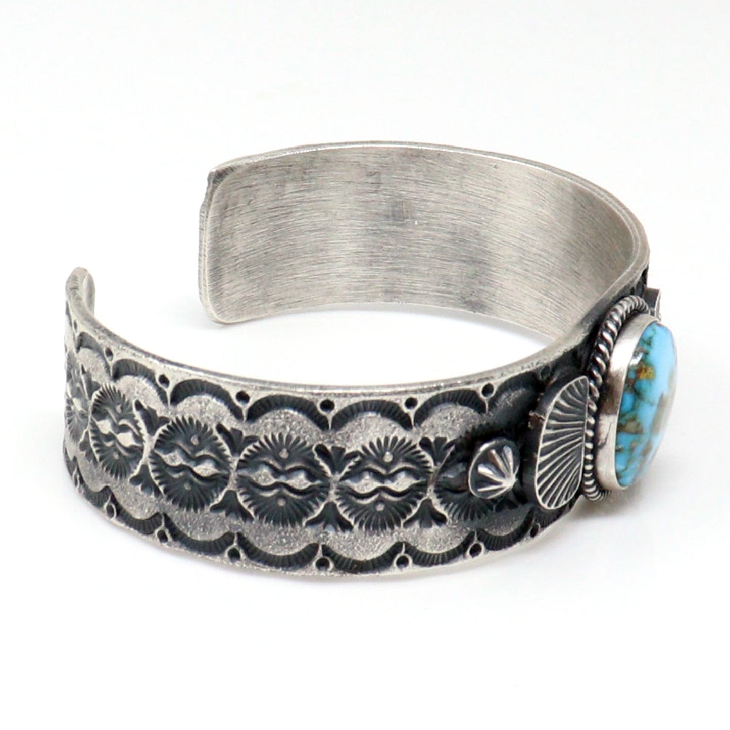 A sterling silver cuff bracelet with hand-stamped floral and sunburst patterns, featuring a round turquoise stone and a small silver conch shell accent on the right side, set against a plain white background.