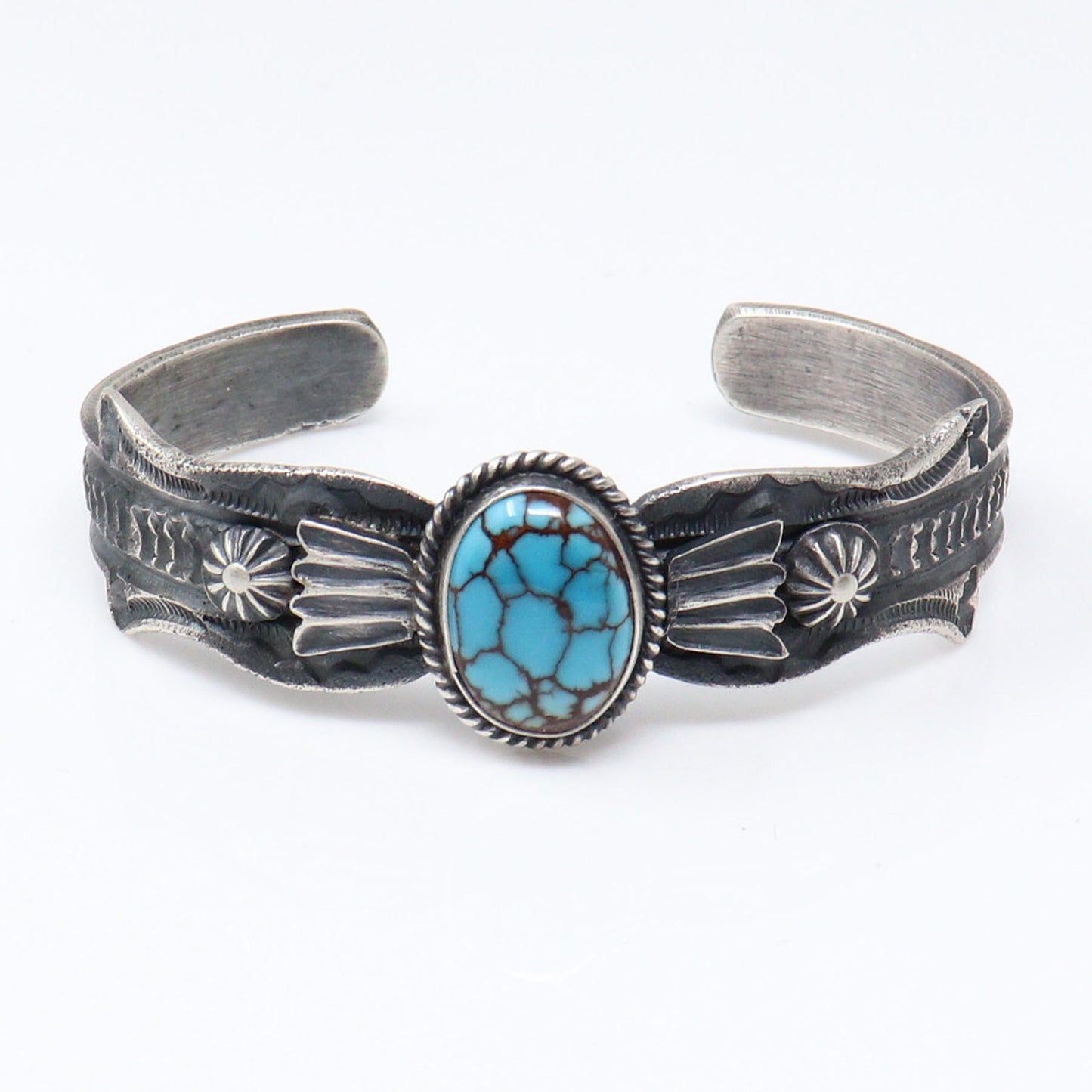 A sterling silver cuff bracelet featuring a central oval turquoise stone with dark matrix veining, surrounded by a braided silver border. The silver band is decorated with hand-hammered details, including sunburst motifs and engraved text, set against a plain white background.