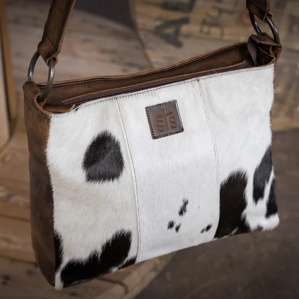 A cowhide shoulder bag with brown leather trim and a brown brand patch on the front, photographed against a rustic wooden background.