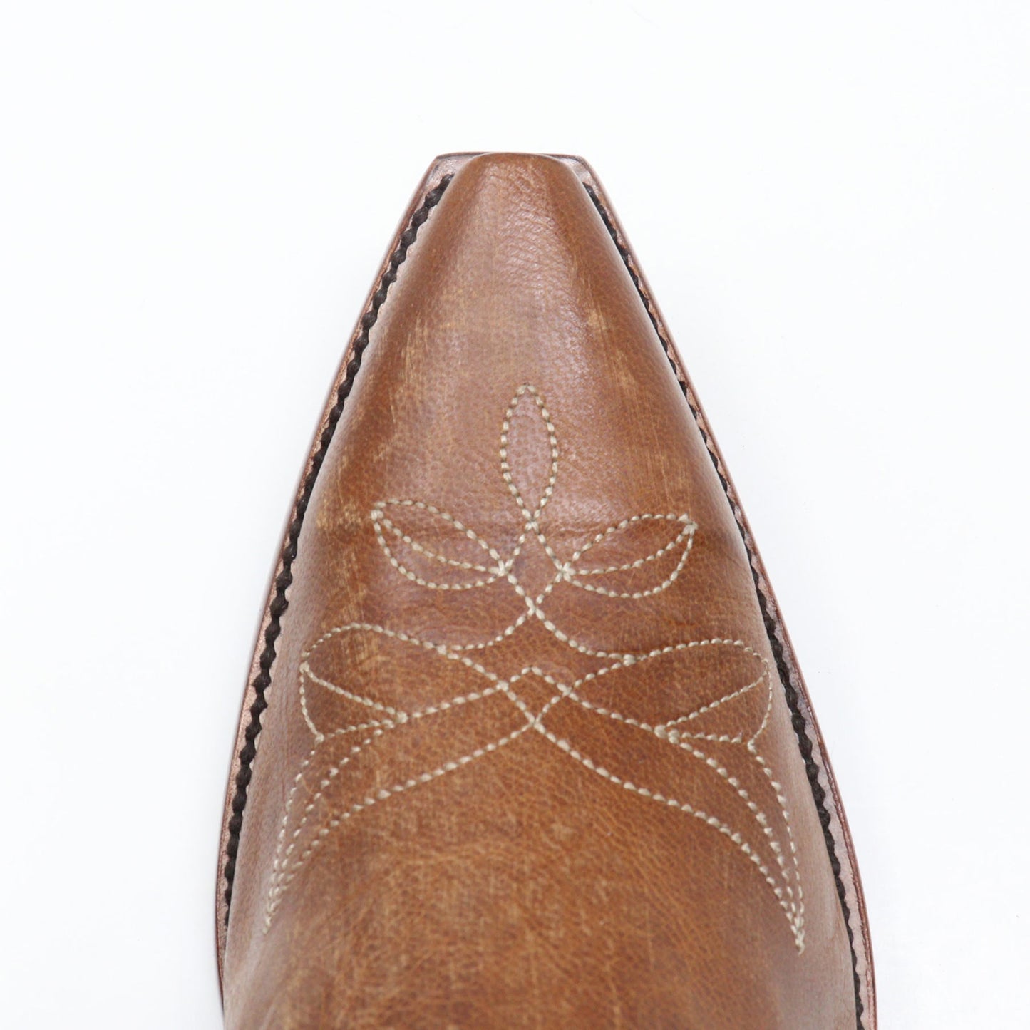 Close-up of a pointed toe on a brown leather boot, featuring decorative white stitching and a textured leather surface on a white background.